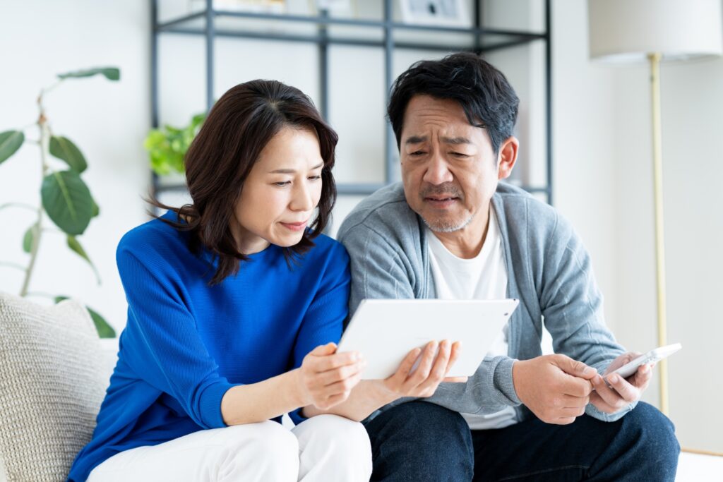 自宅のソファに座り、一緒にタブレットを見ている中年カップル。女性は青いシャツを着て微笑み、グレーのカーディガンを着た男性はスマートフォンを手にタブレットを興味深そうに眺めている。.