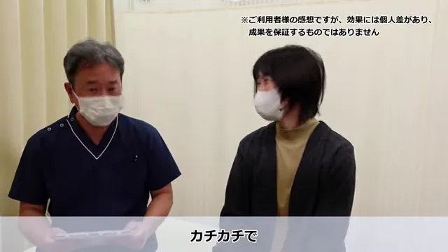 長年の肩こりと繰り返すめまいが根本改善したKさんの声 肩こりがひどく、カチコチの状態を説明する場面。マスク姿の2人が室内で向かい合って座っている。左の人物は黒っぽいスクラブを着ており、右の人物は黒髪でカジュアルな服装。画像の上下に日本語のテキストが表示されている。