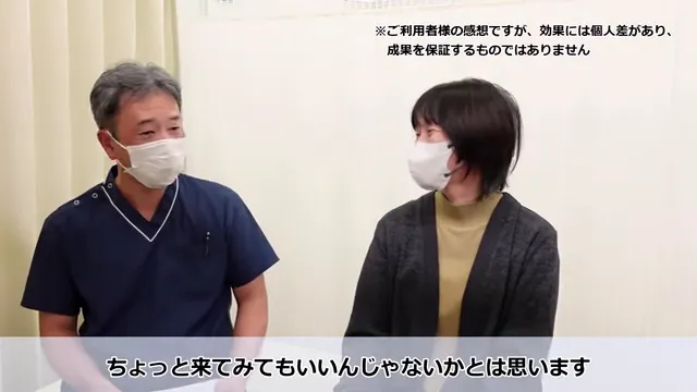 長年の肩こりと繰り返すめまいが根本改善したKさんの声 同じ症状で悩む人へ語りかけるKさんのメッセージシーン。白いマスクをした青いスクラブの男性とベージュのカーディガンの女性が、室内で並んで座り、向かい合って話している。画像の右上と下には日本語のテキストが表示されている。