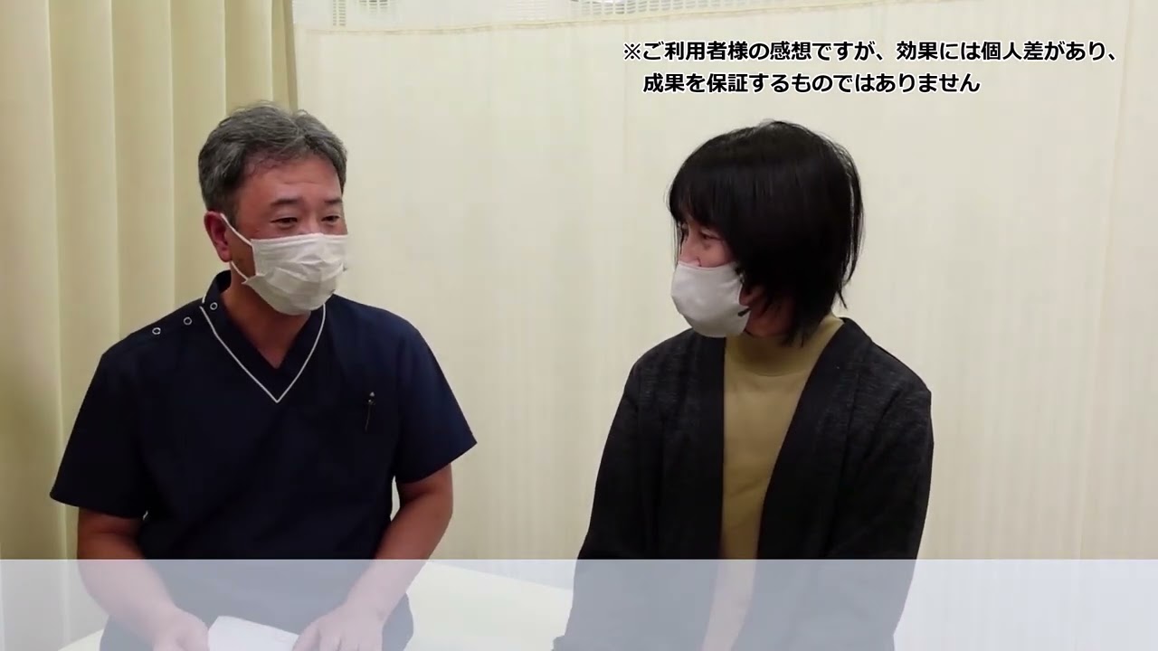 長年の肩こりと繰り返すめまいが根本改善したKさんの声 医療用スクラブ姿の男性とカジュアルな服装の女性が白いマスクをつけて向かい合って座っている。画像右上に日本語の文字が表示されている。